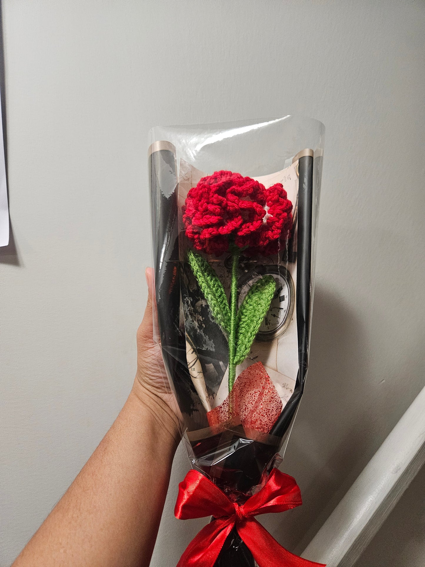Handcrafted crochet red carnation flower with green leaves, wrapped in black and tied with a satin ribbon for a charming, delicate look.