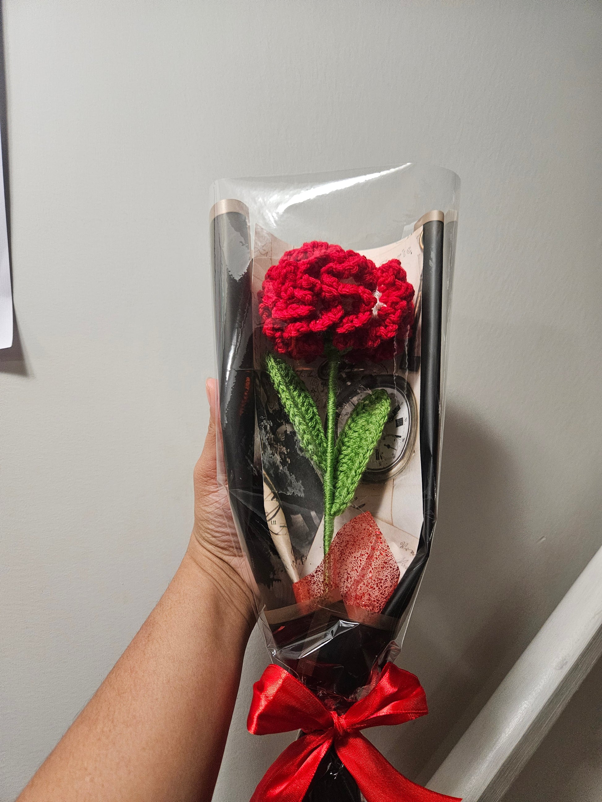 Handcrafted crochet red carnation flower with green leaves, wrapped in black and tied with a satin ribbon for a charming, delicate look.