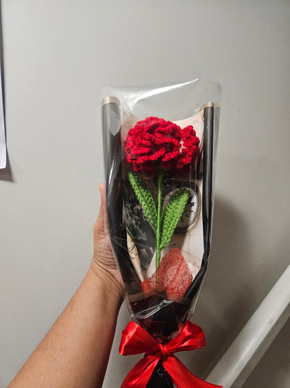 Handcrafted crochet red carnation flower with green leaves, wrapped in black and tied with a satin ribbon for a charming, delicate look.