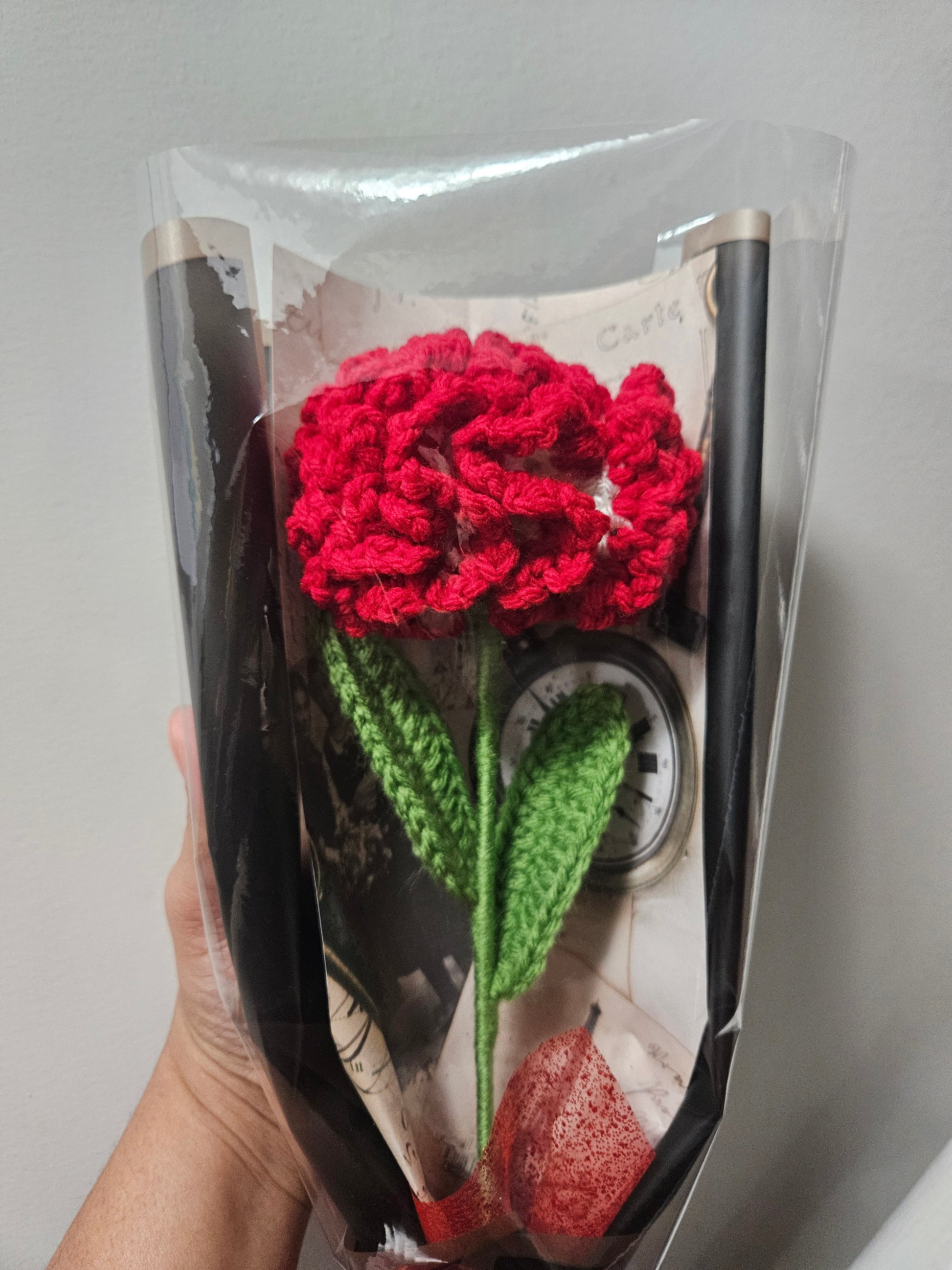 Handcrafted crochet red carnation flower with green leaves, wrapped in black and tied with a satin ribbon for a charming, delicate look.