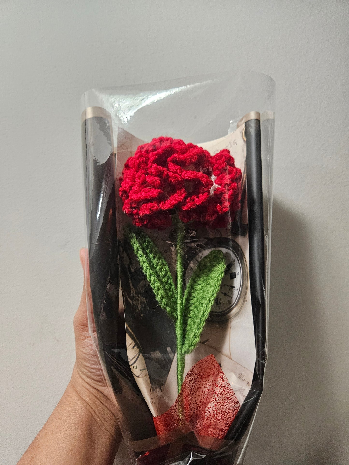 Handcrafted crochet red carnation flower with green leaves, wrapped in black and tied with a satin ribbon for a charming, delicate look.