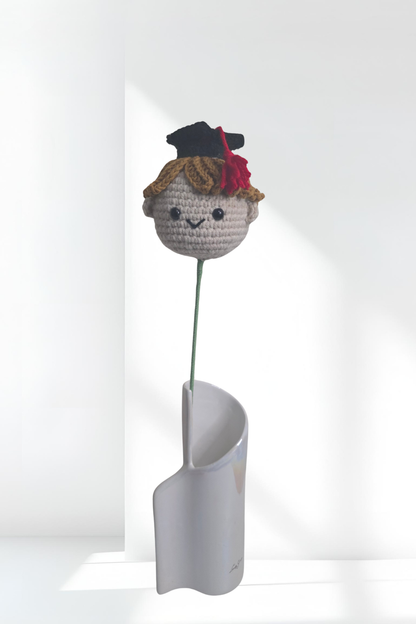 Crocheted character with a graduation cap on a white stand against a white background