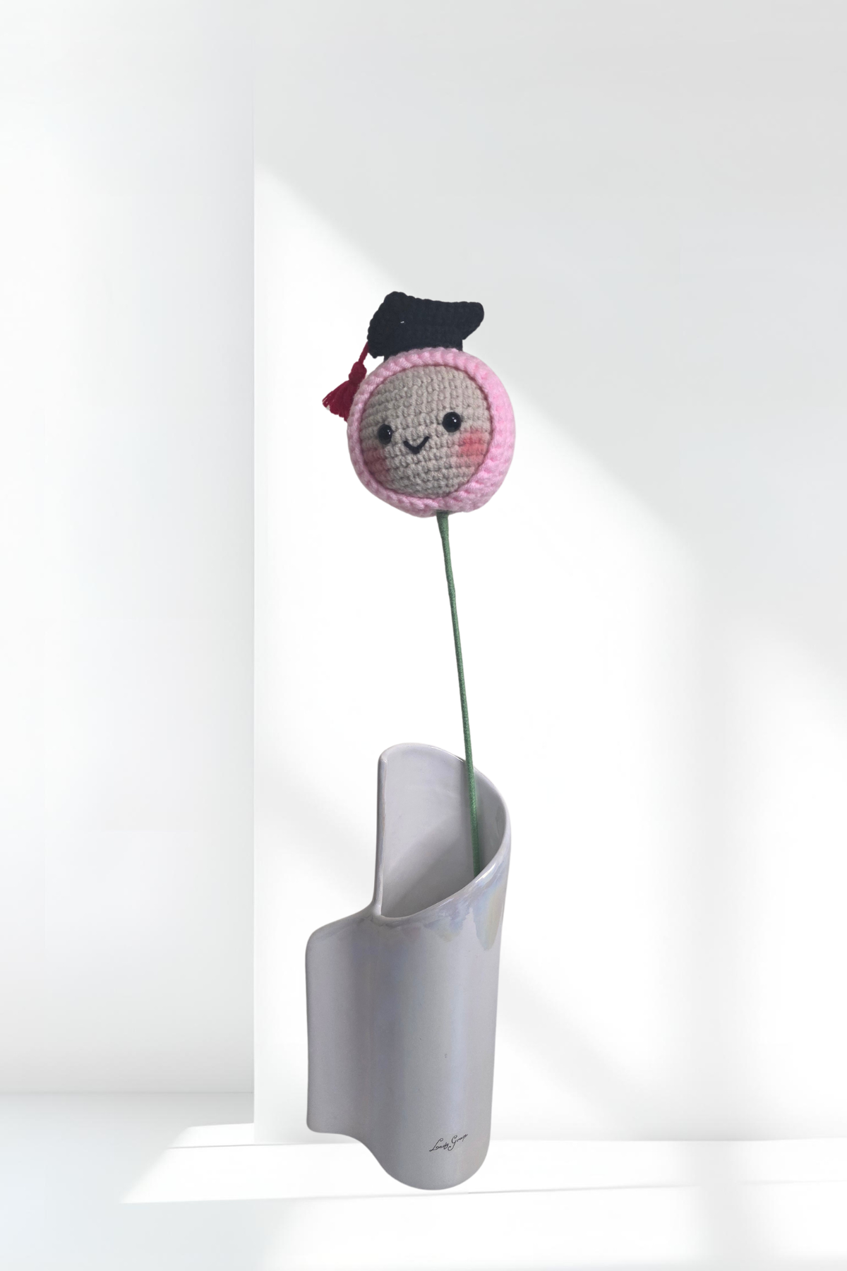 Crocheted flower with a face and graduation cap on a white stand against a white background