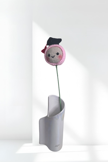 Crocheted flower with a face and graduation cap on a white stand against a white background