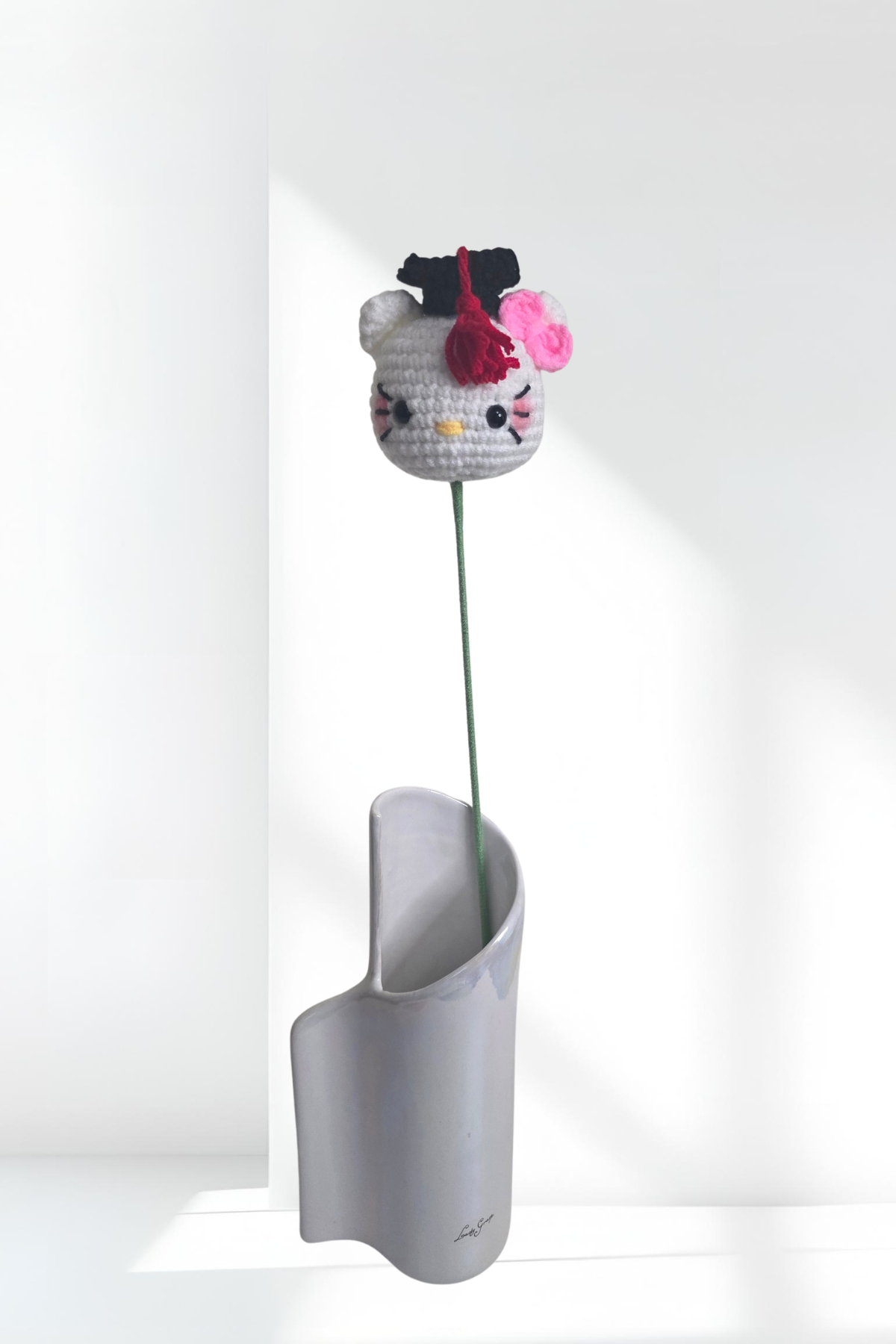 Crocheted Hello Kitty head with a pink flower in a white vase on a white background
