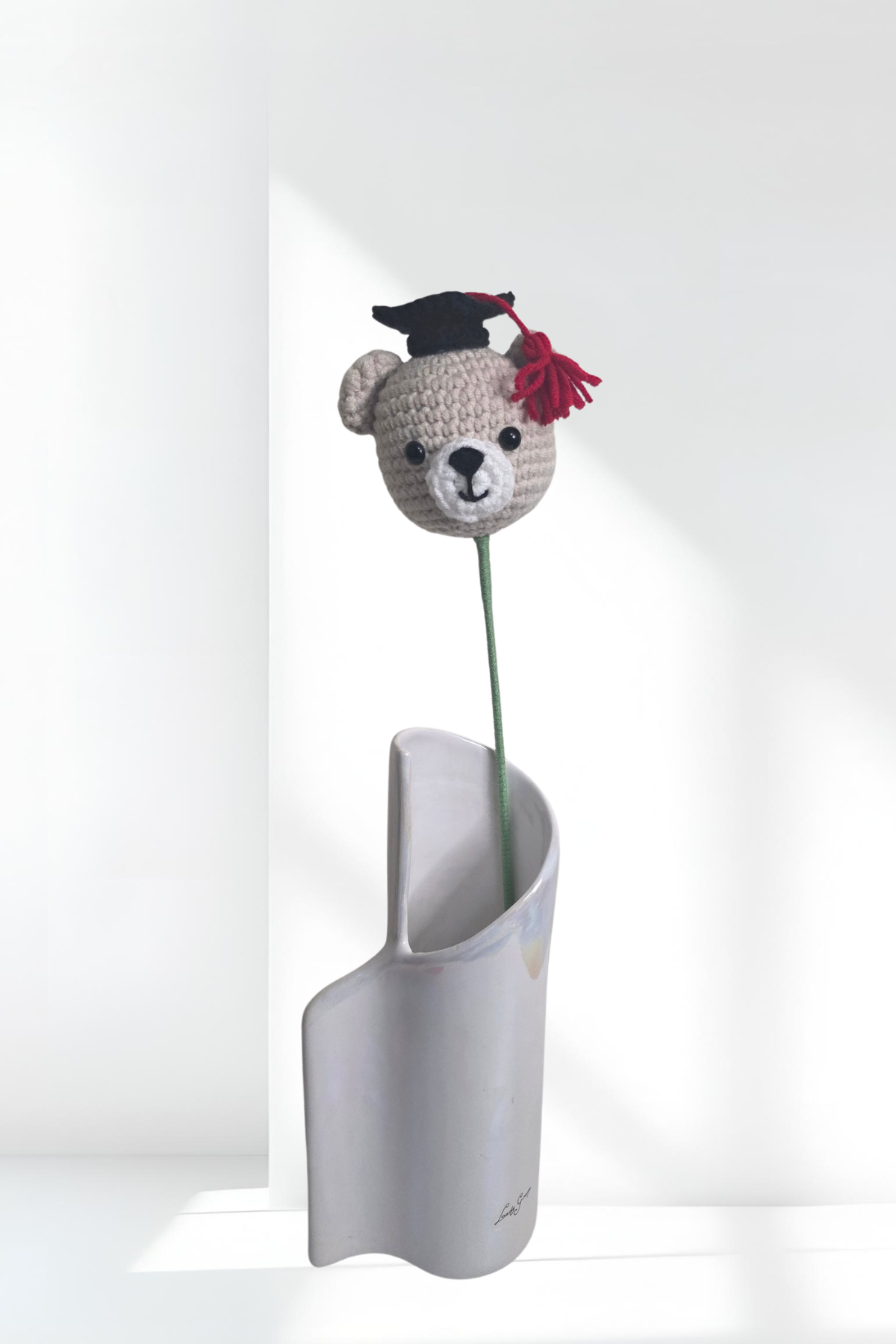 Crocheted bear with a graduation cap and red bow in a white vase on a light gray background