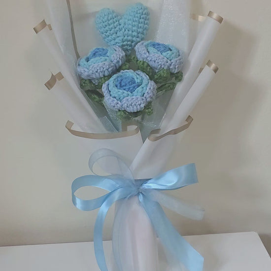 Bouquet of blue crocheted flowers wrapped in white paper with a blue ribbon on a white background